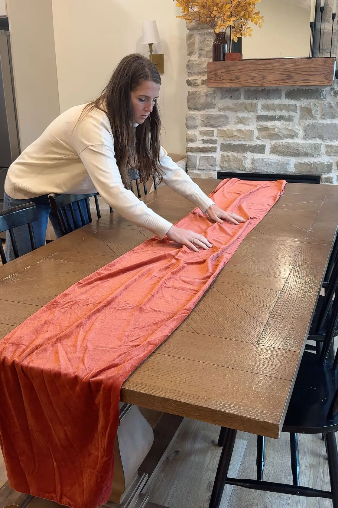 Woman laying an orange velvet table runner across a wooden dining table, setting the foundation for a festive tablescape.